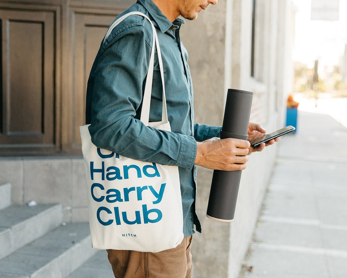 Man holding the Hitch Bottle and Cup in Charcoal Gray stacked in one hand, iPhone in the other hand. One hand carry club tote on his right shoulder.