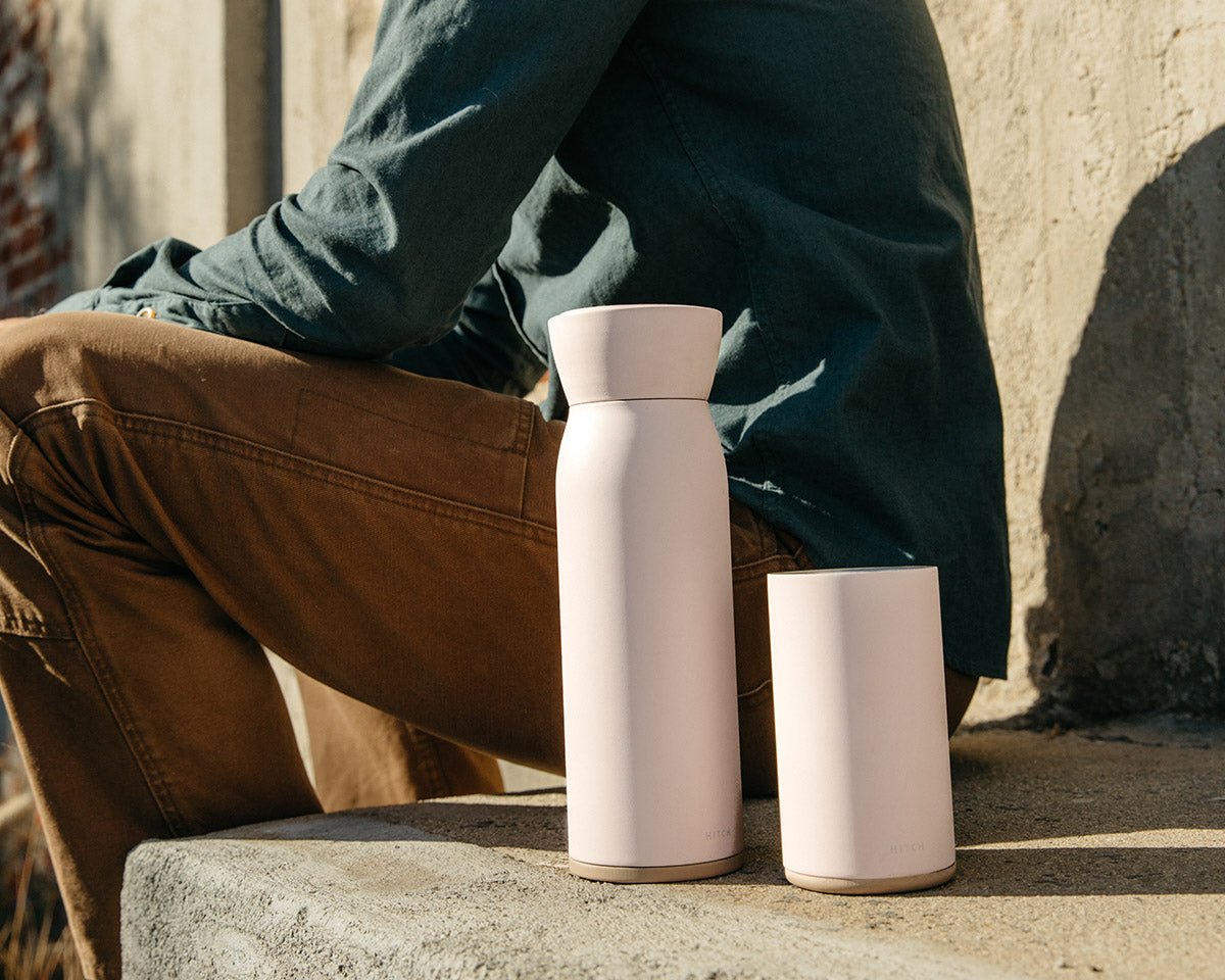 Hitch Bottle and  Hitch Cup in Pale Blush sitting side by side on a concrete stoop next to a seated man
